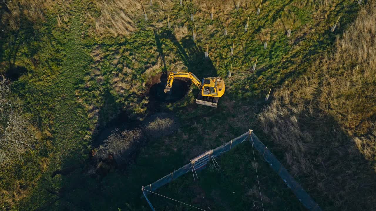 En gravemaskine graver i jorden for at anlægge en dam på en græsbevokset lysning omgivet af træer - luftfoto ovenfra.