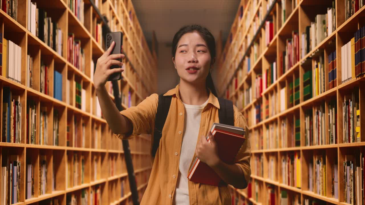 estudiante asiática con una mochila y algunos libros haciendo una llamada de video en un teléfono inteligente mientras está de pie en la biblioteca