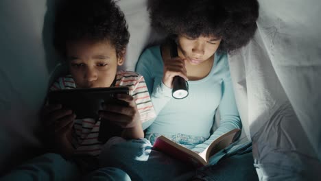 Two-children-spending-night-time-under-blanket-and-reading-book-or-using-phone