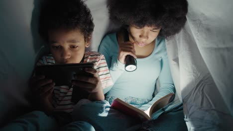 two children spending night time under blanket and reading book or using phone