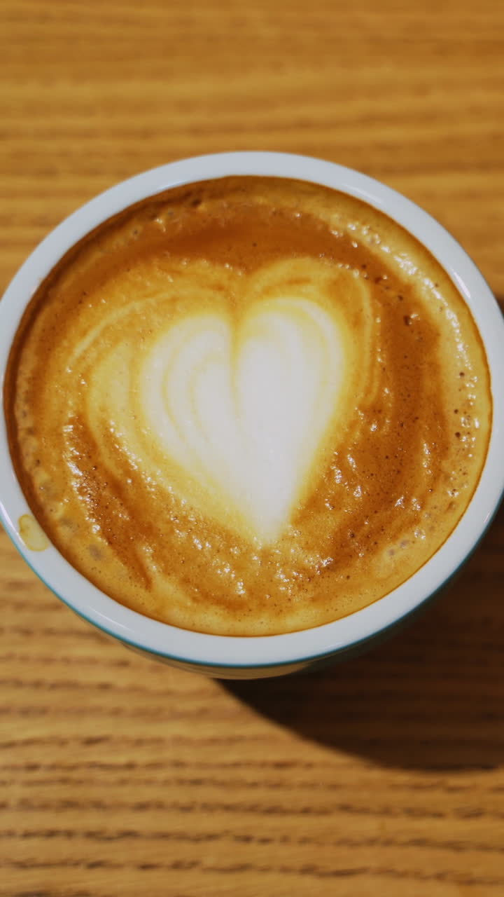 Serving a cup of hot drink. Heart design from milk on coffee. Cup of coffee with latte art in a form of a heart on a wooden background. Close-up. Vertical video