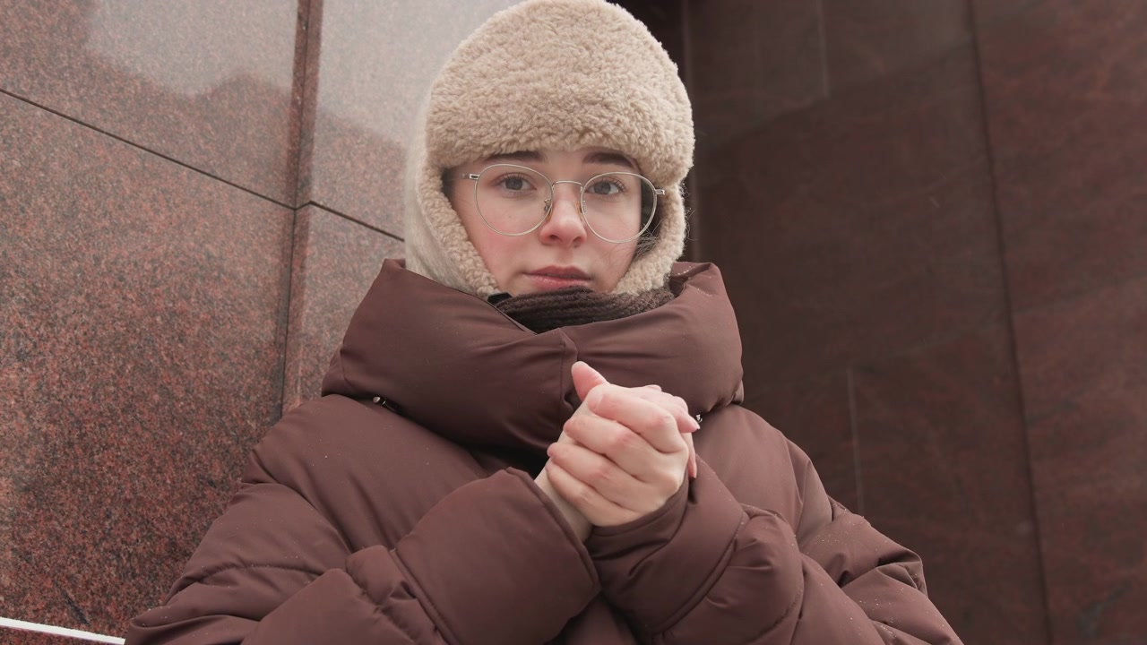 Giovane donna con un pesante piumino marrone e un cappello di pelliccia beige sta accanto a un muro di pietra scura lucida, stringendo delicatamente le mani vicino al petto come per riscaldarsi in una fredda giornata invernale urbana.