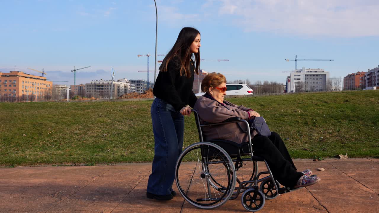 Granddaughter hands phone to grandmother in wheelchair who stores it and gets pushed away for a walk in park