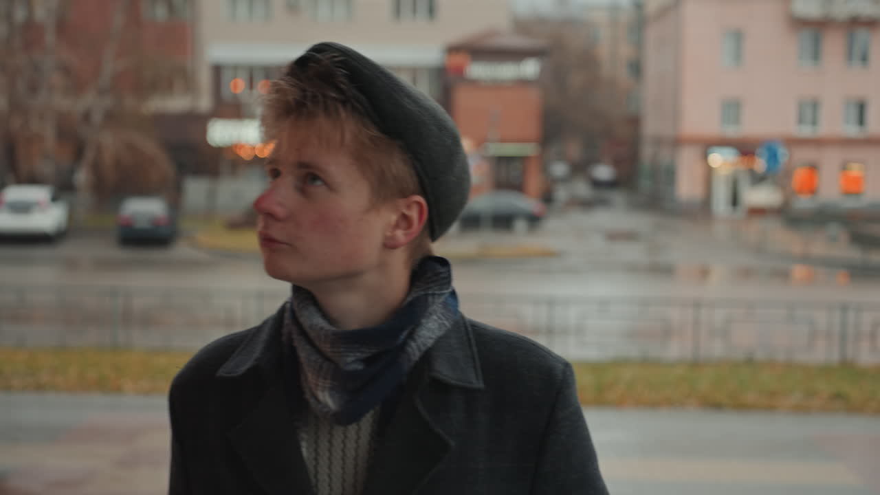 Ung kaukasisk skaber venter under et halvtag, regnfuld bygade, hænder i frakkelommer, scanner butiksfacader, fjern trafik-bokeh, melankolsk holdning, poetisk stemning, diskret bevægelse antyder