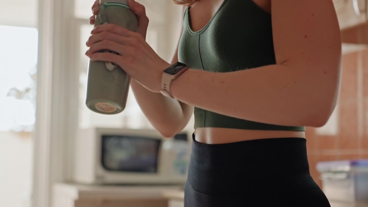 Donna in cucina post-allenamento che agita una bottiglia di proteine. Primo piano del busto con reggiseno sportivo verde e smartwatch, mani che afferrano lo shaker, luce brillante dalla finestra, polvere di integratore mescolata nella bevanda, per un recupero rapido.