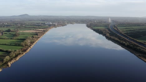 Antena:-Río-Suave-Y-Vidrioso-Refleja-El-Cielo-Y-Las-Nubes,-Puente-Distante