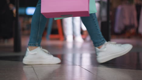 lower leg view of someone in jeans and canvas sneakers carrying a mint and pink shopping bag in their right hand while walking past a clothing store in a mall