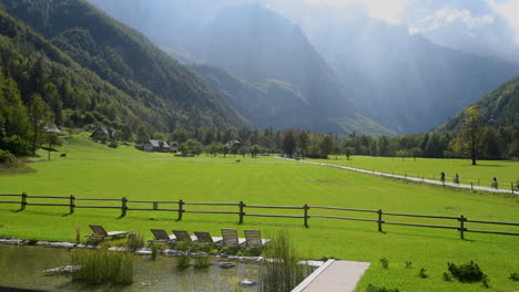 panoramic view in logarska valley, slovenia, green meadows with forest and high mountains in background, natural swimming pool with sunchairs in front