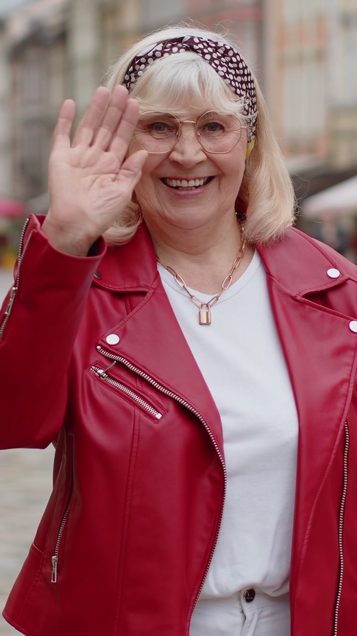 Donna anziana che sorride amichevolmente alla telecamera agitando le mani ciao ciao saluto o addio in strada della città