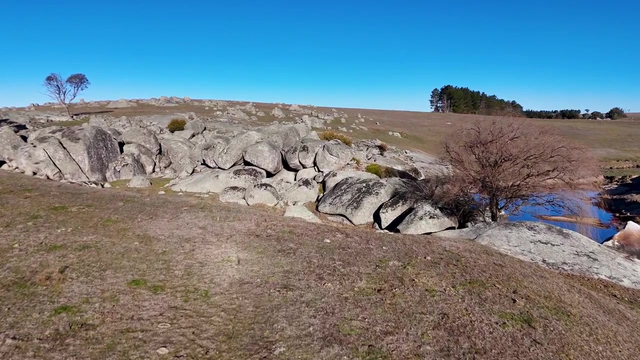 Drone panoroi graniittisten kalliomuodostelmien ja pienen puron yli kirkkaan sinisen taivaan alla.