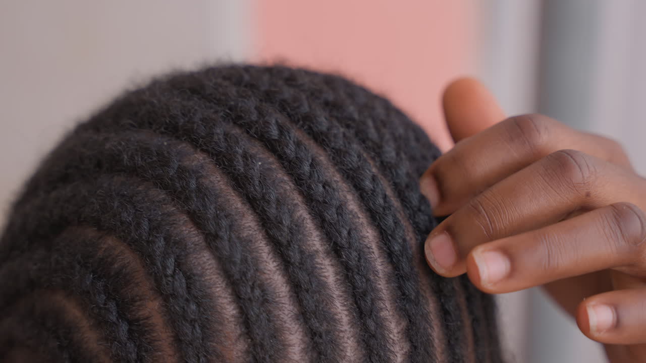 Gros plan sur un enfant noir dont les tresses plaquées sont ajustées, la main de la mère lissant délicatement les rangées nettes contre un mur rose doux, lumière naturelle, détail du cuir chevelu, cheveux texturés, routine intime de soins capillaires à domicile, protectrice.