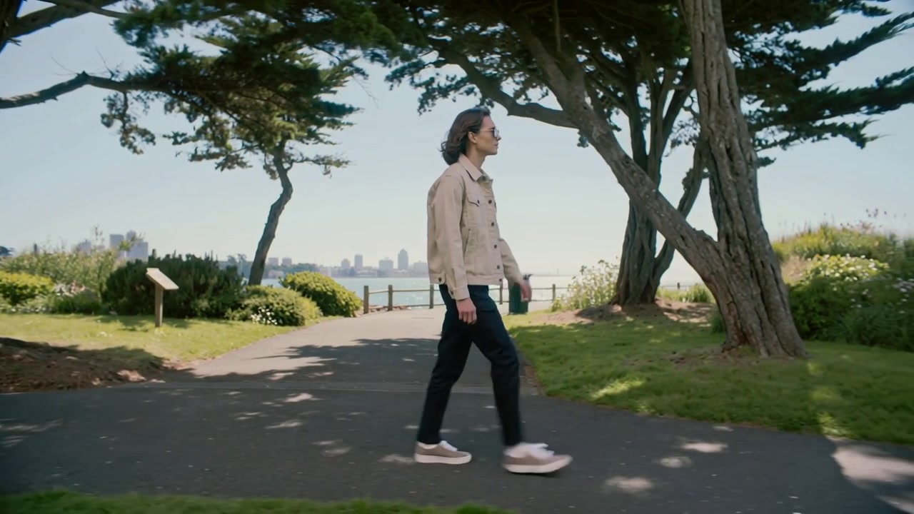 Walking man starting mid-stride, crossing waterfront path under trees, in tan jacket and sunglasses