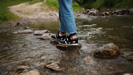 Shooting-close-up-a-girl-in-jeans-and-black-and-white-sneakers-crosses-a-mountain-river-along-a-special-path-from-stones