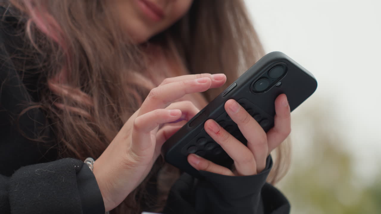 Giovane studentessa caucasica che usa lo smartphone all'aperto, primo piano delle mani che digitano e scorrono su una cover nera, capelli lunghi e cappotto invernale visibili, controllo dei messaggi e composizione di appunti per la lezione, studio spontaneo