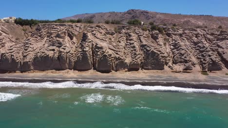 aerial flying sideways alongside greek island, rocky cliff and blue water waves in the mediterranean sea on beach in santorini, greece