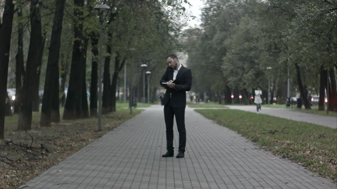 joven hombre de negocios en el callejón del parque hablando por teléfono y escribiendo en el diario, trabajando con notas en el parque verde