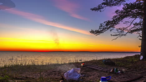 Two-friends-enjoy-a-peaceful-picnic-on-a-tranquil-beach,-as-a-vibrant-sunset-paints-the-sky-in-shades-of-orange-and-purple