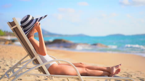 Sexy-Mujer-Exótica-En-Traje-De-Baño-Y-Sombrero-Disquete-Disfrutando-En-La-Silla-De-Playa-Junto-Al-Mar-Tropical-En-El-Soleado-Día-De-Verano,-Cámara-Lenta-De-Marco-Completo