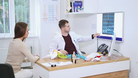 Doctor shows the x-ray to a female patient. Young woman listening to a specialist in modern clinic. Spine model on the table. Health care concept.