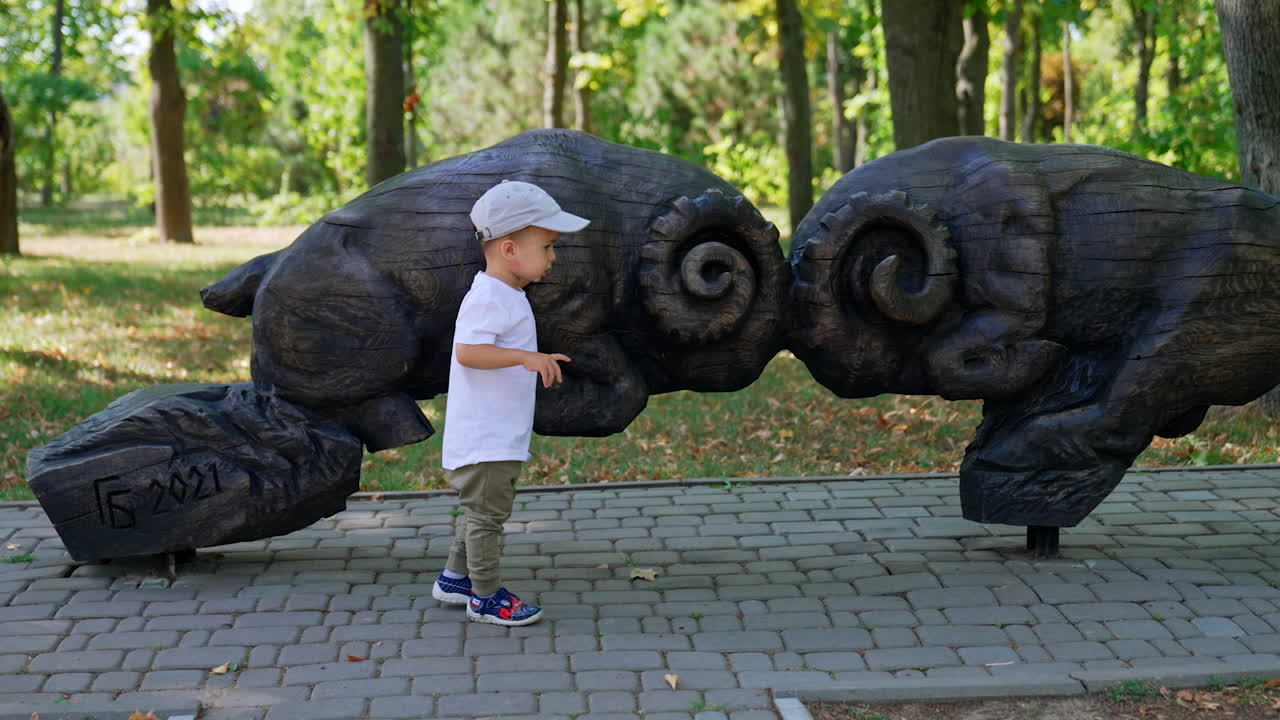 Pensive toddler standing at the black wooden sculptures of sheep in the park. Child touches the figures and walks around them.