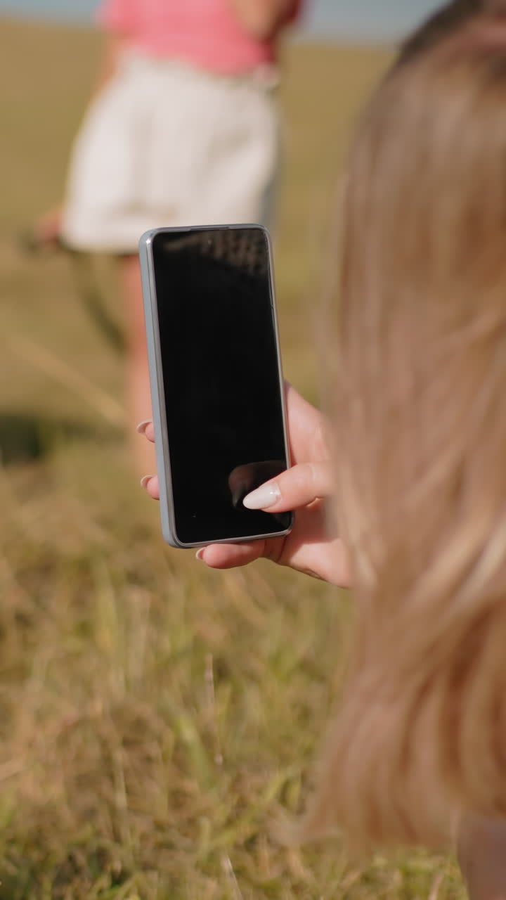 vista trasera de cerca de una mujer tomando una foto de una persona acostada en el heno con las piernas hacia arriba, rodeada de un campo verde exuberante, vista borrosa de dos niños jugando en el fondo