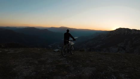 Joven-Parado-Con-Su-Bicicleta-En-La-Cima-De-Una-Montaña-Al-Atardecer