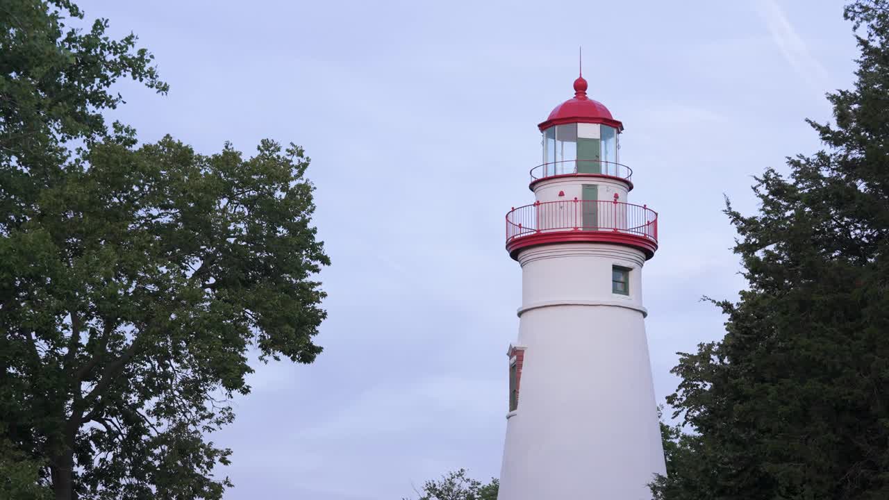 A still midshot of the Marblehead Lighthouse in Port Clinton, Ohio, framed by trees and clear sky, highlighting its striking red and white architectural details