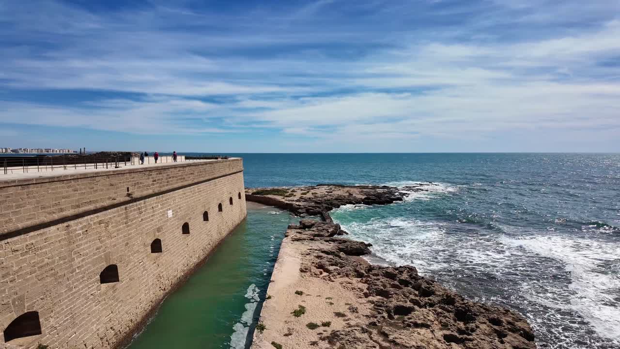 İspanya'nın Cádiz şehrindeki tarihi deniz duvarının yanındaki kayalık kıyıya çarpan dalgalar. Taş tahkimat, Atlantik kıyısı boyunca parlak mavi bir gökyüzünün altında uzanarak denizcilik tarihini yansıtıyor.