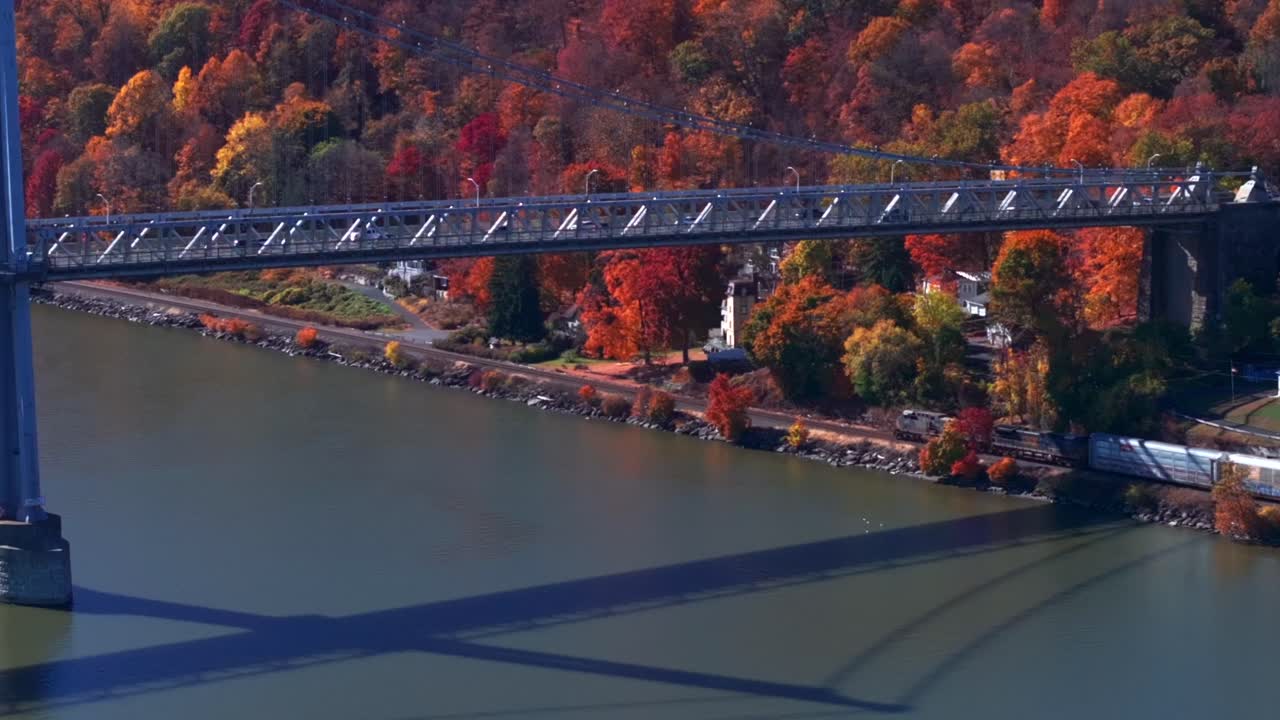 Aerial view over the Hudson River next to the Mid-Hudson Bridge on a sunny day in autumn. The camera dolly in and boom down following a cargo train traveling under the bridge.