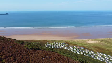 retreating aerial shot over moorland view of rhossili bay with caravan holiday park and worm's head landmark with winding path trails on sunny summer day 4k