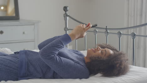 Smiling-Young-Woman-Lying-On-Bed-With-Mobile-Phone-Wearing-Pyjamas