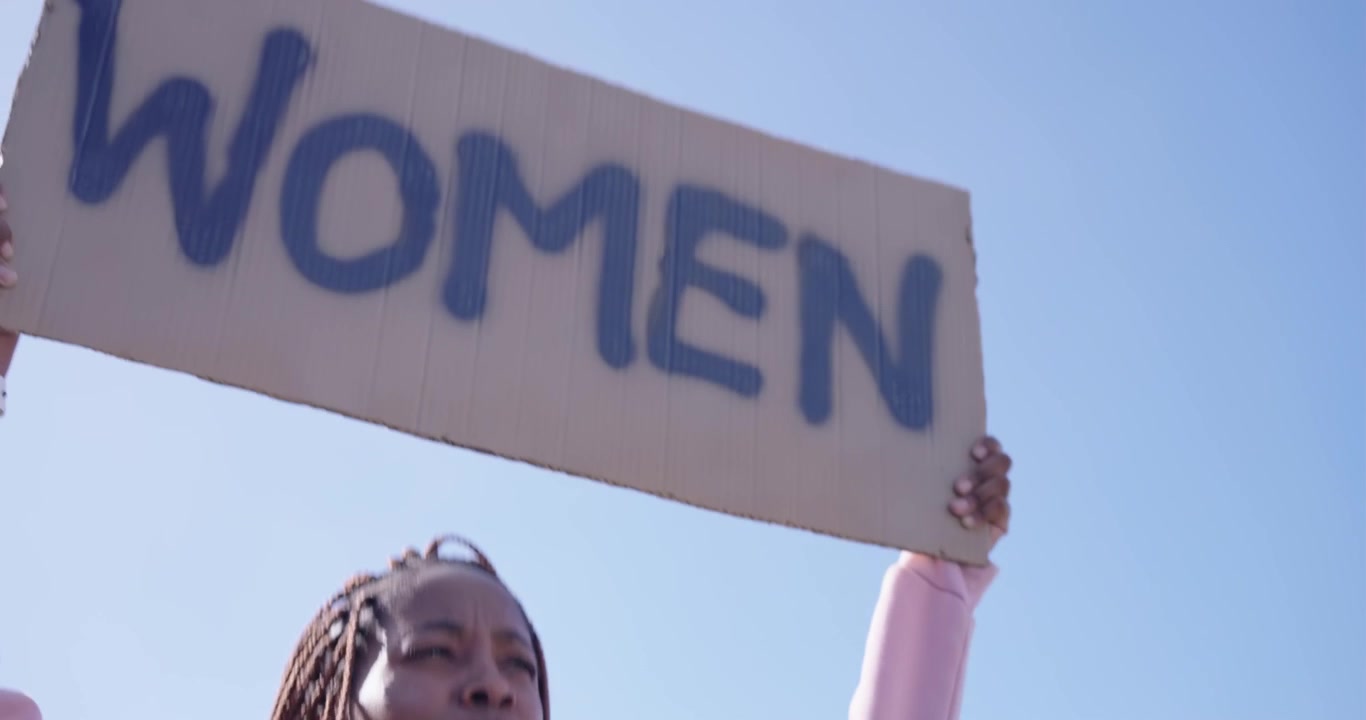 Afroamerikanische Frau in pinkem Kapuzenpullover auf einem Dach, die Arme erhebend, skandierend und ein „WOMEN“-Schild für eine Kundgebung haltend.