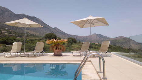 gimbal shot pushing in on sun loungers on patio of luxury greek villa with pool in foreground and mountains in background