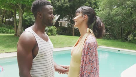 Happy-diverse-couple-holding-hands-at-swimming-pool-in-garden