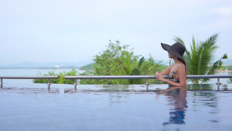 elegant woman with large back hat bathing and relaxing in infinity pool overlooking the ocean