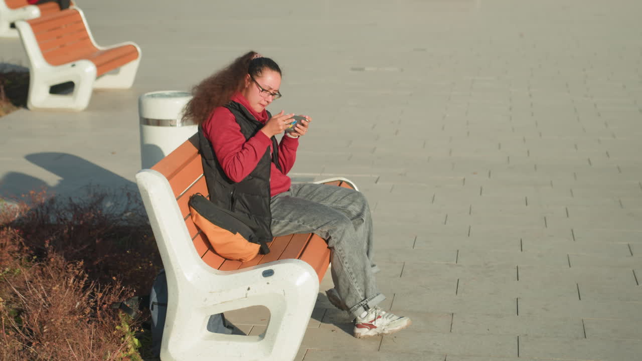 静かな都会の公園のベンチでスマホをスクロールする若い女性、赤いジャケットとグレーのジーンズ、隣にバックパック、写真やメッセージを確認中、暖かい日差しと長い影、自然なライフスタイルの一瞬、秋