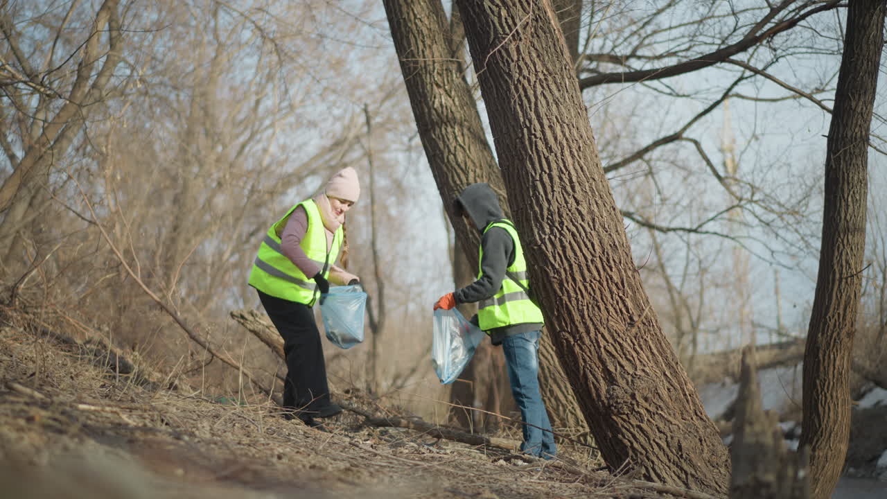 Two volunteers wearing reflective safety vests and gloves picking up litter along riverbank during outdoor cleanup, placing trash into blue bags to support environmental protection