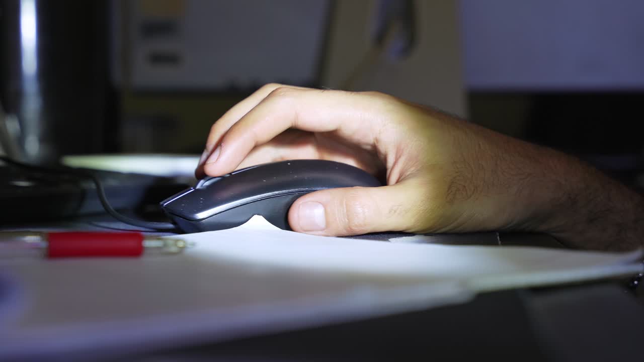 Una mano che usa un mouse nero per computer su una scrivania di notte. Scena cinematografica in condizioni di scarsa illuminazione con una penna rossa in primo piano sfocato, ideale per temi di lavoro a distanza o studio notturno.