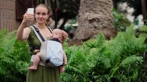 accompanied by her baby in a kangaroo backpack, a young woman documents her journey with a mobile phone. as she walks, she checks the phone screen