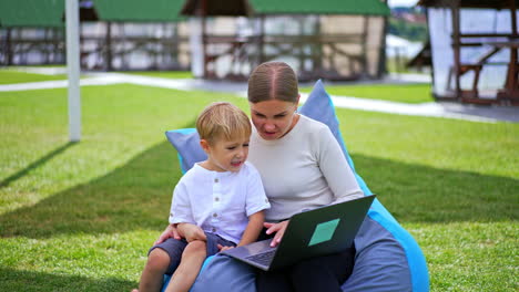 mom and son of five years old sitting close in one bean bag chair. family spending time together outside looking at computer.