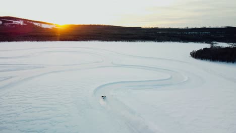 toma de drone de un auto deportivo conduciendo en un lago congelado durante la puesta de sol con puesta de sol y montañas al fondo en el círculo ártico