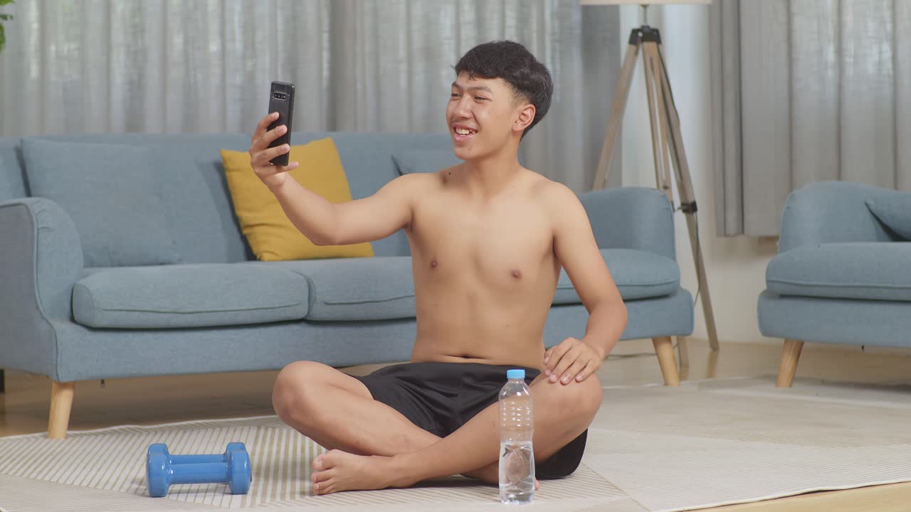 아시아 십대 소년 은 집 에서 셔츠 를 벗고 운동 한 후 덤벨 을 들고 매트 에 앉아 스마트폰 으로 비디오 통화를 하는 동안 손 을 흔들고 있다