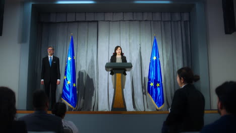 female politician makes an announcement, answers journalists questions and gives interview for media. confident representative of the european union during press conference. backdrop with eu flags.