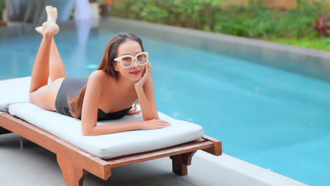 young asian girl in black monokini lying on her belly on a comfortable deck chair in a gorgeous tropical resort poolside smiling, daytime slow-motion miami