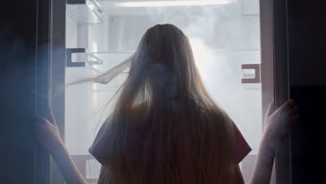 vista trasera de una mujer joven mirando dentro del refrigerador. el viento y la niebla soplan en su cara y mecen su cabello