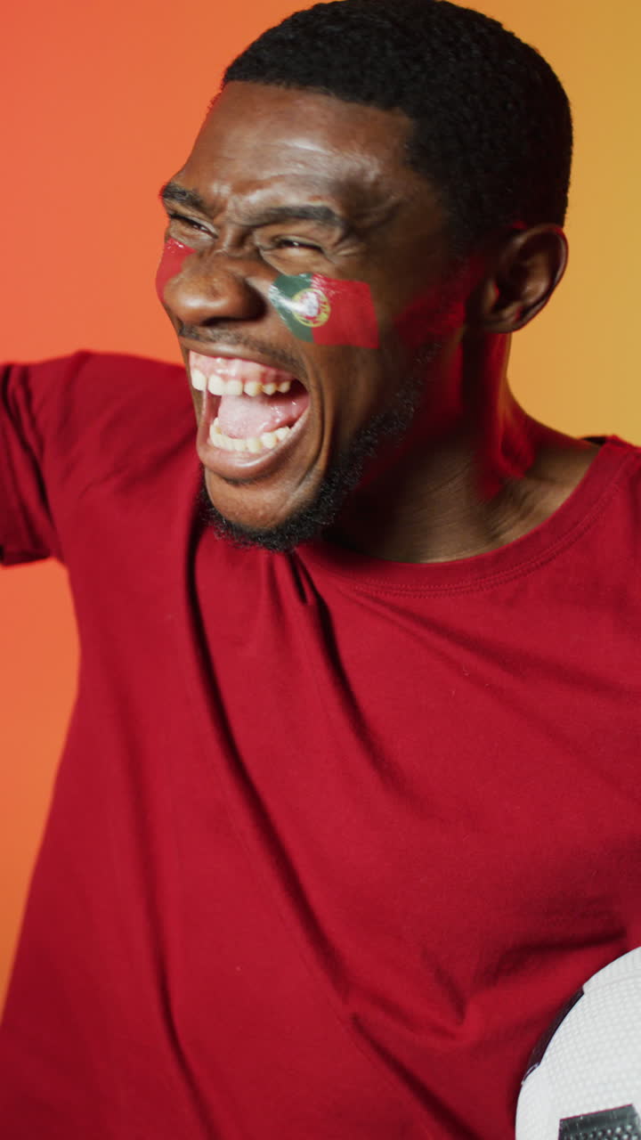 Happy african american male football supporter with flag of portugal on cheeks over pink lighting