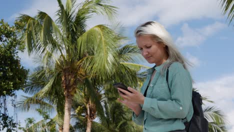 beautiful slim woman with long blonde hair in green shirt standing near palm tree and using smartphone over background the park. girl on the square touching screen and smile