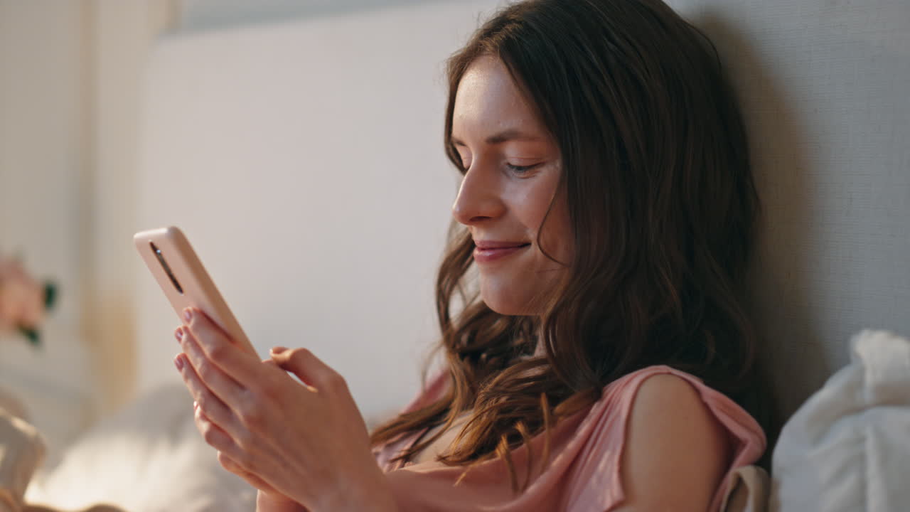 Primer plano de mujer tranquila bebiendo café por la mañana en la cama. Chica sonriente enviando mensajes en línea.