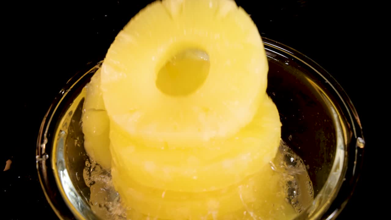 A pineapple ring falls into a clear glass bowl, creating a water splash. High-speed close-up, black background, bright studio lighting, minimalistic composition