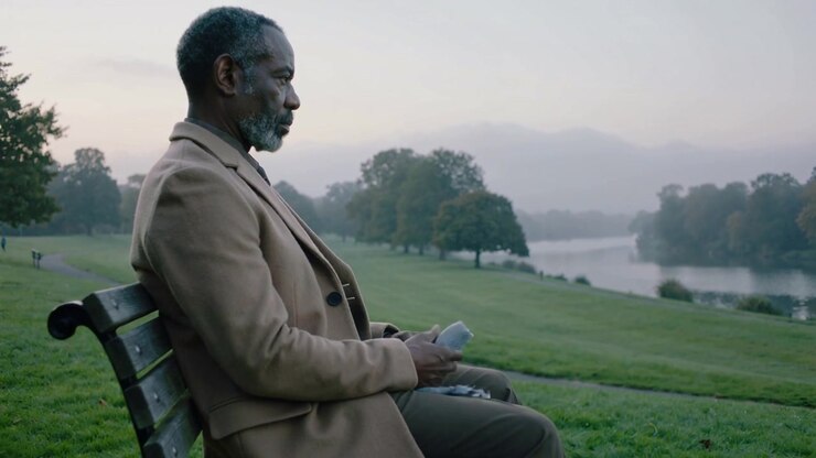 Reacting to dawn haze, man on park bench wearing camel coat adjusting scarf, holding cup